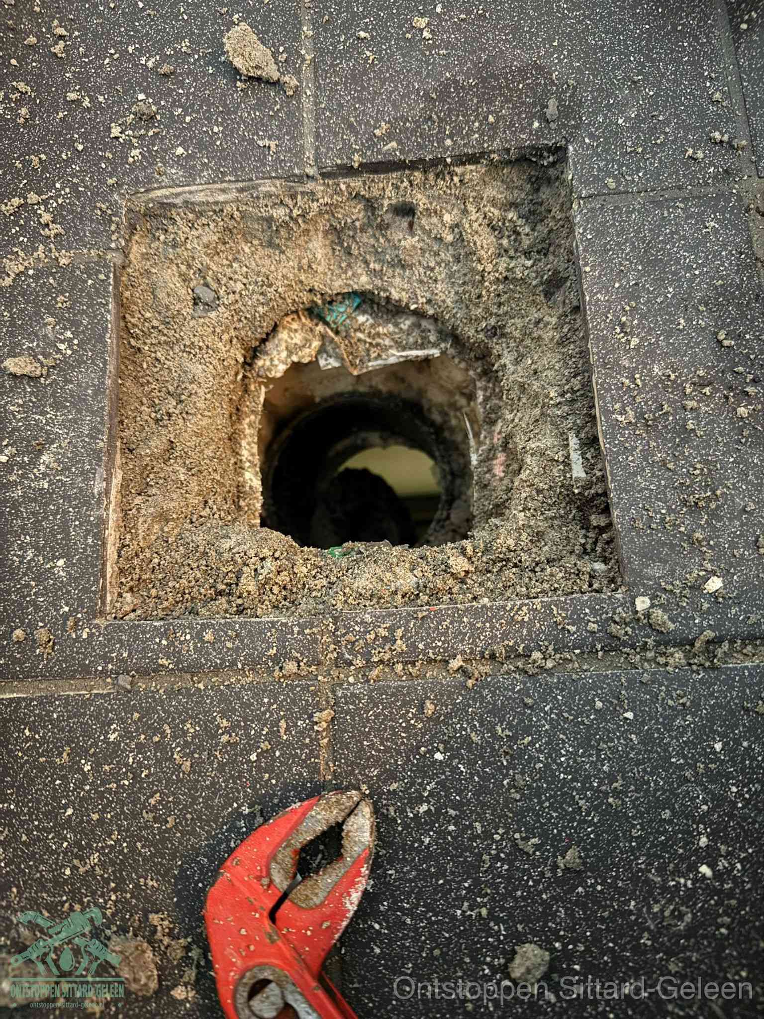 Een geopend rioolputje in een betontegelvloer met losgemaakt rooster en een zichtbare ronde afvoerbuis, het achterhalen waar de afvoerverstopping zit in Sittard-Geleen