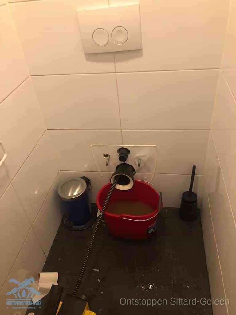 Een ontstopte wand-wc met verwijderde toiletpot waar een ontstoppingsveer in de wanduitgang verdwijnt, het achterhalen waar de verstopping zit in Sittard-Geleen