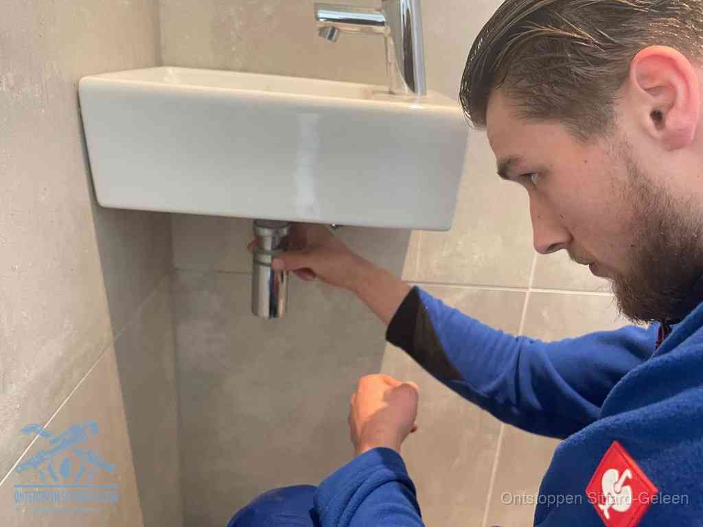 Een monteur draait met de hand de zwanenhalssifon onder een fonteinwasbak in de toiletruimte los om de bron van rioollucht in Sittard-Geleen te vinden