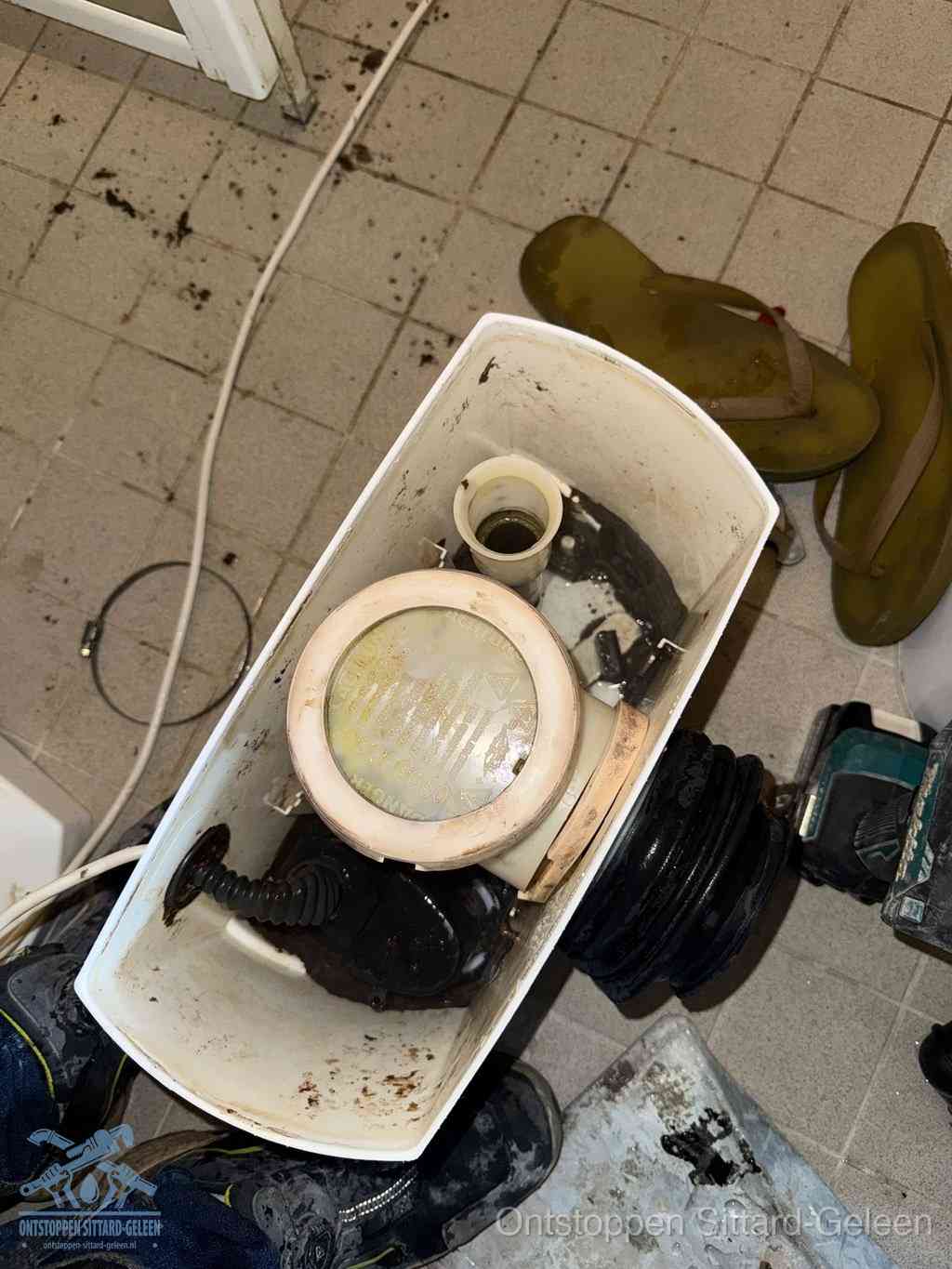 Een gedemonteerde toiletpot op de vloer met ontstoppingsveer en gereedschap ernaast, het verwijderen van een vastzittend voorwerp uit de wc in Sittard-Geleen